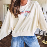 Cropped creamy light weight crew pullover with long sleeves and American flag embroidery with a light blue bow on the front.