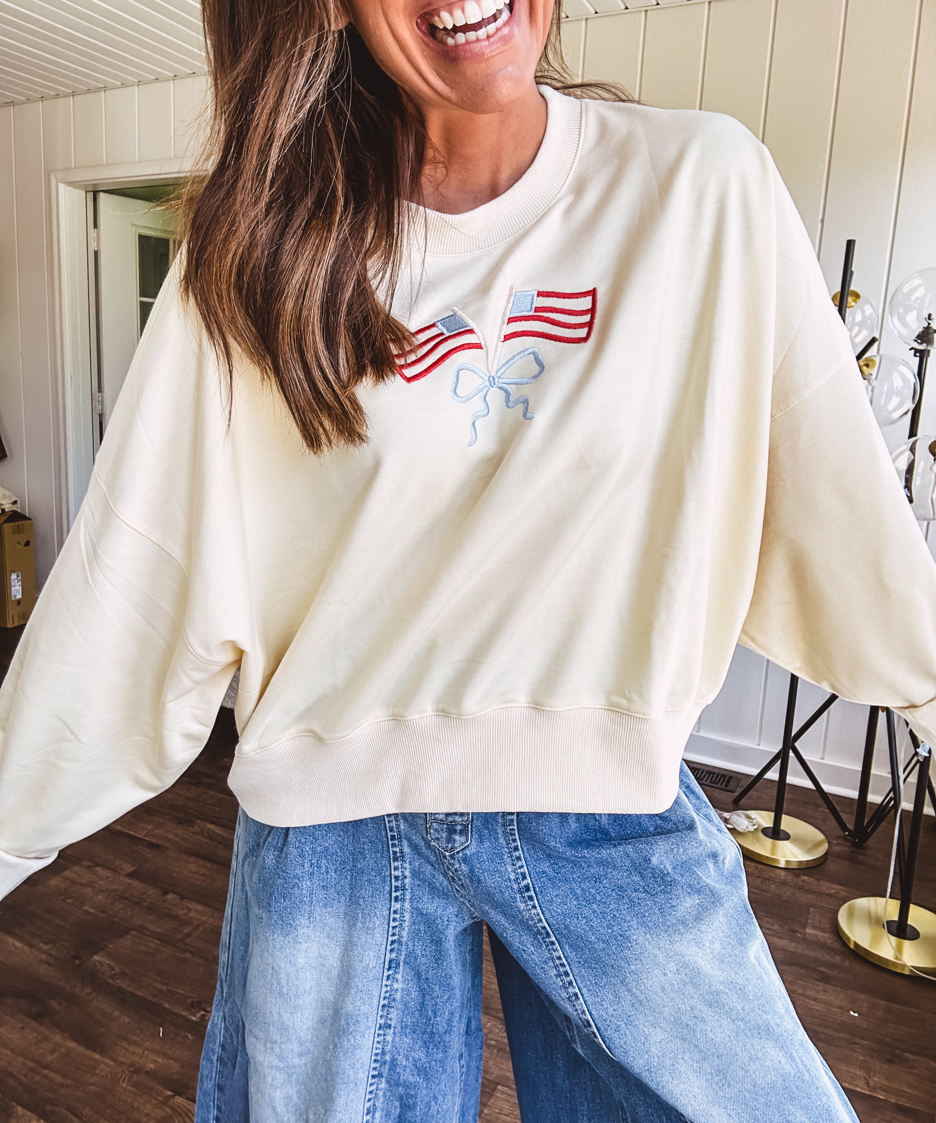 Cropped creamy light weight crew pullover with long sleeves and American flag embroidery with a light blue bow on the front.