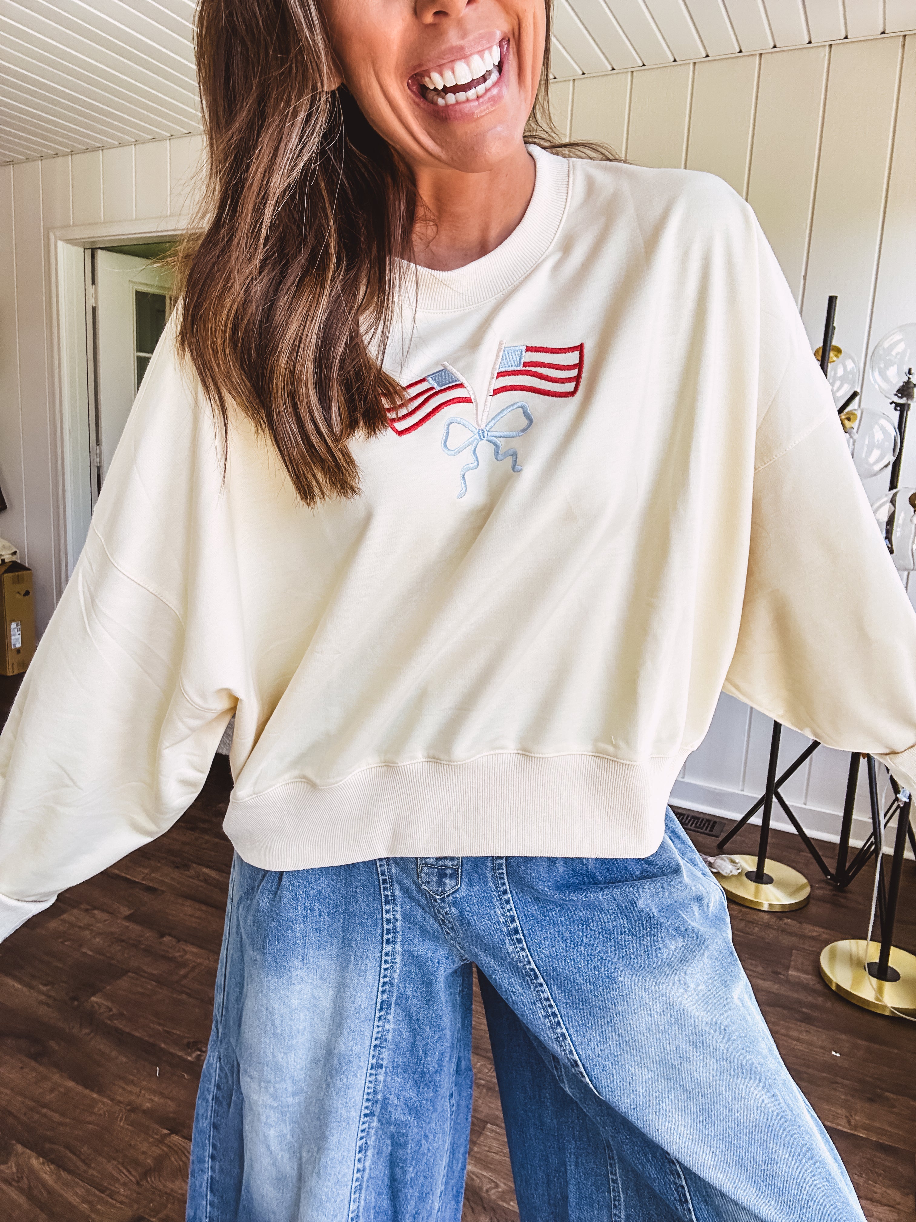 Cropped creamy light weight crew pullover with long sleeves and American flag embroidery with a light blue bow on the front.