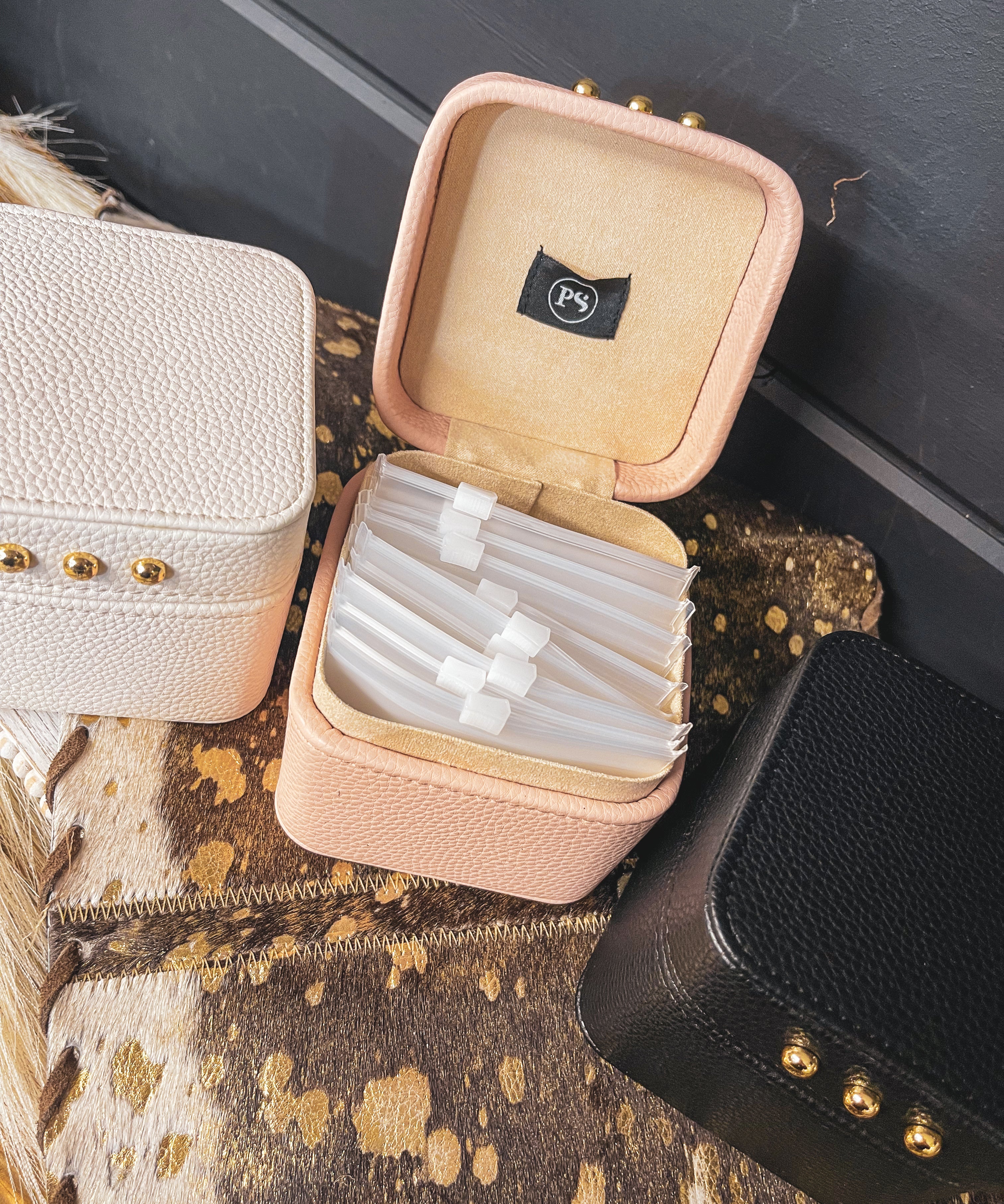 Square leather jewelry travel box with magnetic closure, individual transparent plastic zip closure bags inside and three gold balls on the front. Available in white, pink or black.