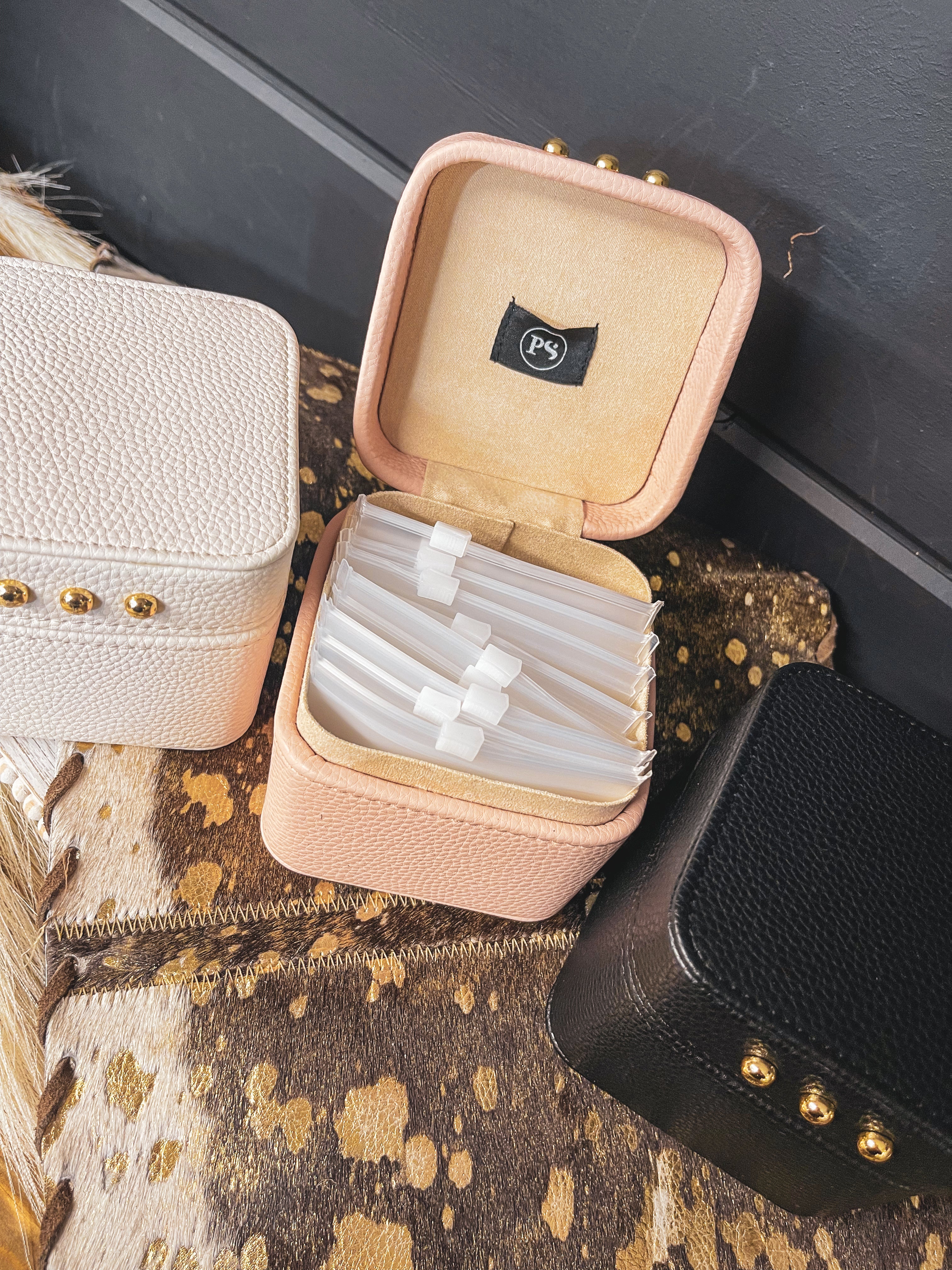 Square leather jewelry travel box with magnetic closure, individual transparent plastic zip closure bags inside and three gold balls on the front. Available in white, pink or black.