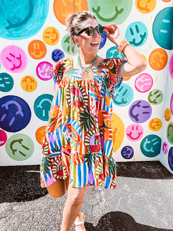This tropical print short dress features vibrant summer hues with playful striping and fruit motifs throughout, square smocked neckline and flutter sleeves and pockets—complete with pink and blue ball tassels. Crafted from soft, lightweight fabric, it's designed to move with you all day long.