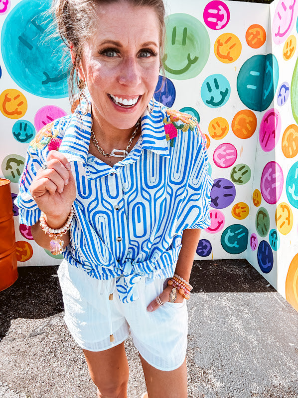 Light blue and white geometric short sleeved button down with collar and cuffed sleeves with bold orange, green and pink embroidery on the shoulders.
