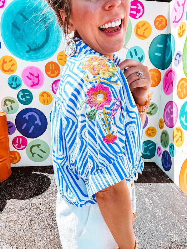 Light blue and white geometric short sleeved button down with collar and cuffed sleeves with bold orange, green and pink embroidery on the shoulders.