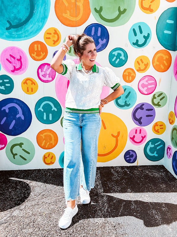 Nearly sheer ivory eyelet patterned blouse with a collar, puff sleeves and three large rhinestone floral buttons at the neckline. The collar, cuffs and hem are a contrasting bright green with pink athletic striping.