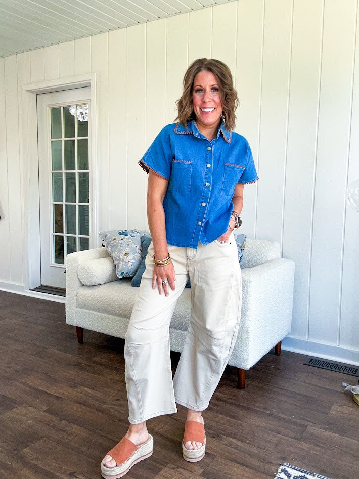Medium denim blue short sleeved top with collar and snap button closure with two front pockets and brown trim at the collar, pockets and cuffs.