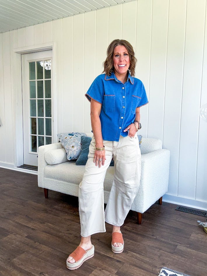 Medium denim blue short sleeved top with collar and snap button closure with two front pockets and brown trim at the collar, pockets and cuffs.