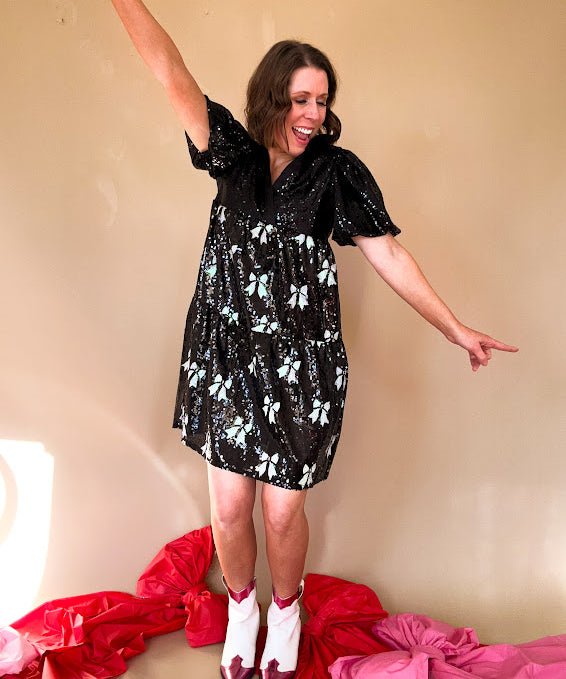 Black sequined mini dress with a silk V-neckline and balloon sleeves, an empire waist with white sequined bows on the skirt. 