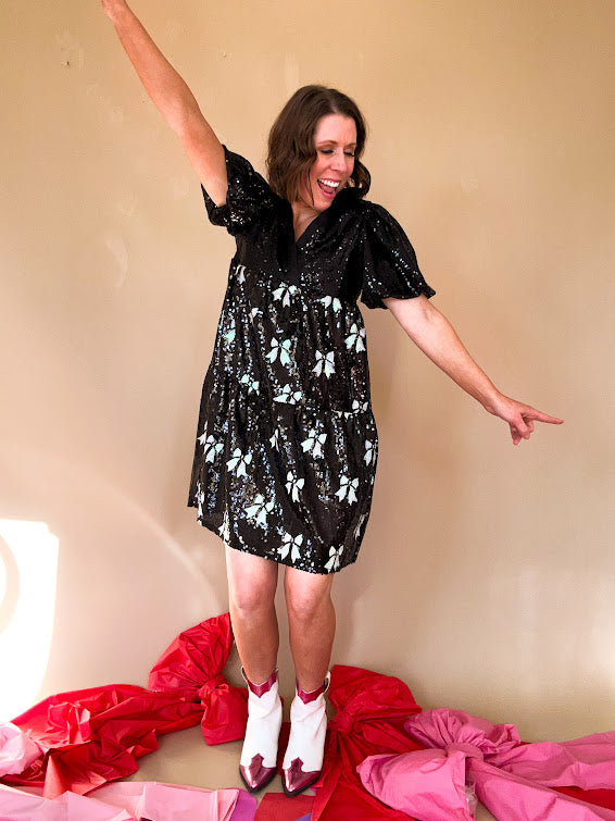 Black sequined mini dress with a silk V-neckline and balloon sleeves, an empire waist with white sequined bows on the skirt. 