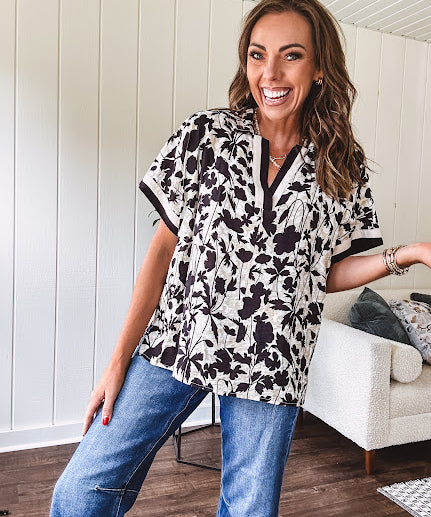 Thin, lightweight black and cream tropical print drop shoulder blouse with cream and black striping at the V neck, and short sleeve cuffs, standard collar and split flowy hem.