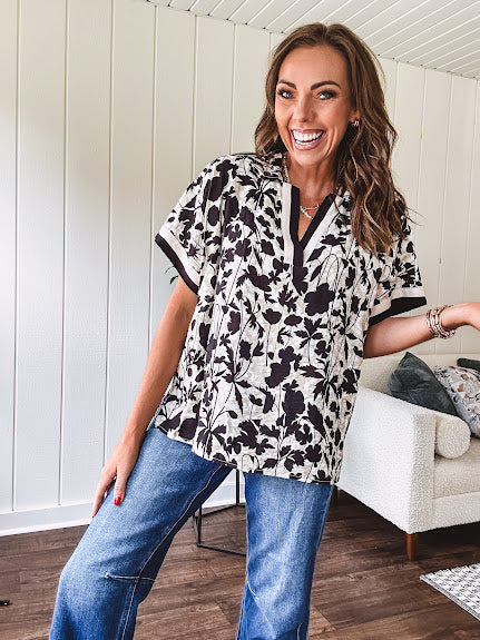 Thin, lightweight black and cream tropical print drop shoulder blouse with cream and black striping at the V neck, and short sleeve cuffs, standard collar and split flowy hem.