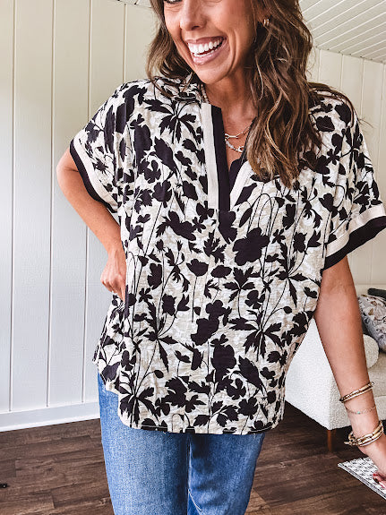 Thin, lightweight black and cream tropical print drop shoulder blouse with cream and black striping at the V neck, and short sleeve cuffs, standard collar and split flowy hem.
