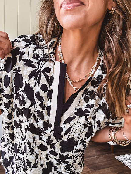 Thin, lightweight black and cream tropical print drop shoulder blouse with cream and black striping at the V neck, and short sleeve cuffs, standard collar and split flowy hem.