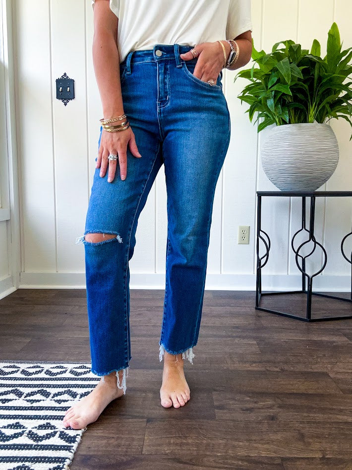 High rise dark wash denim with button and zipper closure, stretch, distressing at the knee and raw hem with straight leg.
