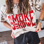 100% Cotton  tee with bold red "Smoke Show" graphic against an artistic tan and brown paint splatter backdrop.