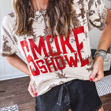 100% Cotton  tee with bold red "Smoke Show" graphic against an artistic tan and brown paint splatter backdrop.