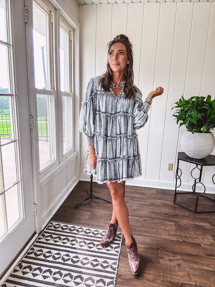 Soft, lightweight Tencel denim mini dress in a retro acid wash design with a V neck and mock collar and tiered design with long sleeves and elastic cuffs each tier and the mock neck feature raw seams and it has pockets.