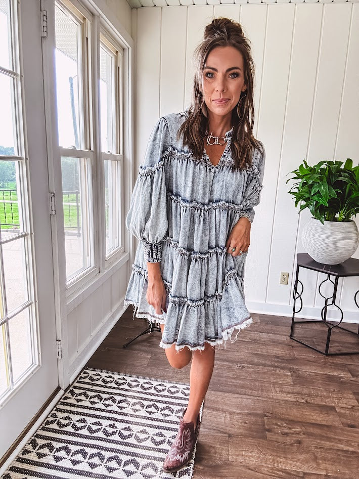 Soft, lightweight Tencel denim mini dress in a retro acid wash design with a V neck and mock collar and tiered design with long sleeves and elastic cuffs each tier and the mock neck feature raw seams and it has pockets.