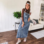 Medium wash denim dress with spaghetti straps and silver metal adjusters, a unique button and zipper front resembling the top of a pair of jeans, back pockets, side pockets, a back slit and raw hem.