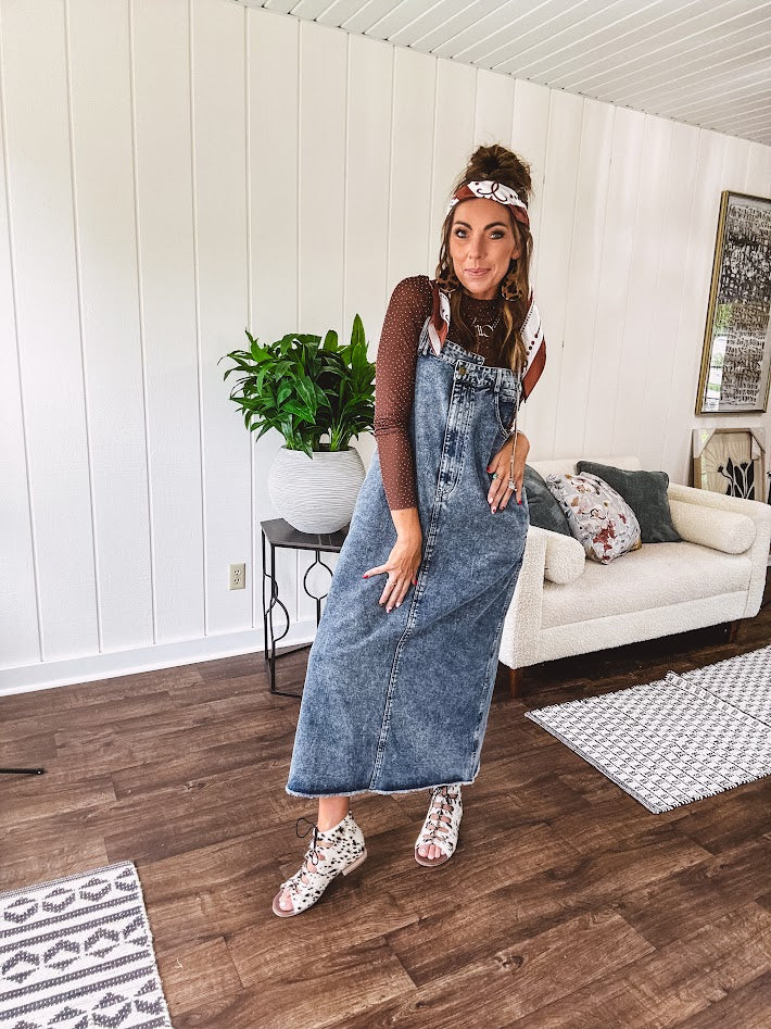 Medium wash denim dress with spaghetti straps and silver metal adjusters, a unique button and zipper front resembling the top of a pair of jeans, back pockets, side pockets, a back slit and raw hem.