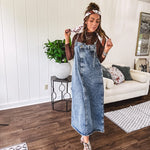 Medium wash denim dress with spaghetti straps and silver metal adjusters, a unique button and zipper front resembling the top of a pair of jeans, back pockets, side pockets, a back slit and raw hem.