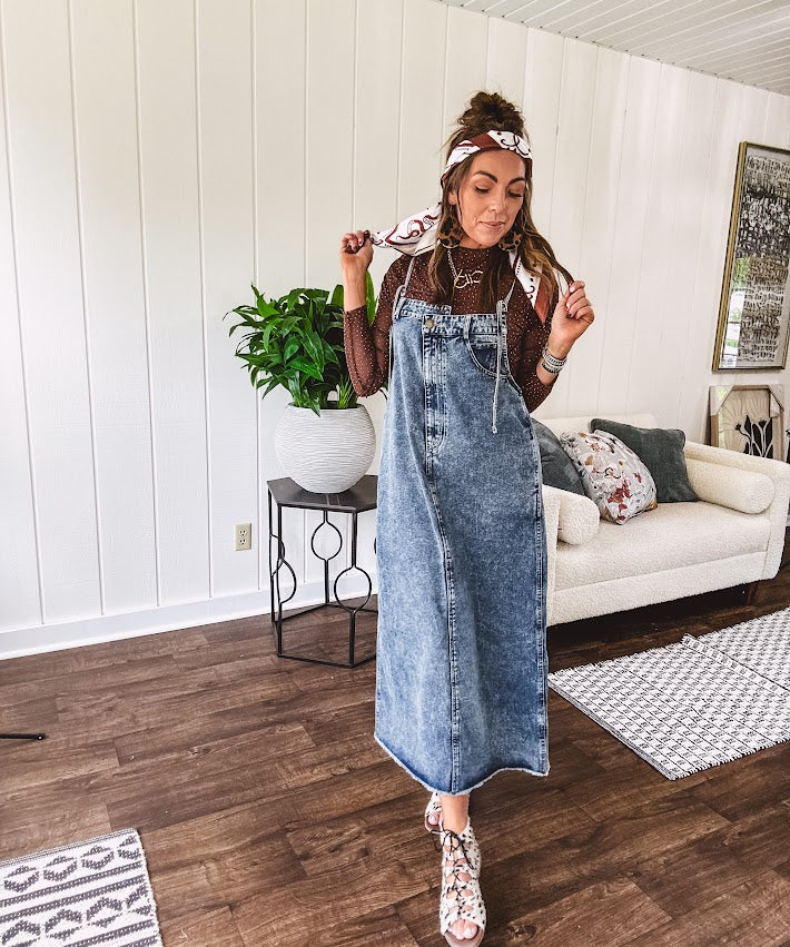 Medium wash denim dress with spaghetti straps and silver metal adjusters, a unique button and zipper front resembling the top of a pair of jeans, back pockets, side pockets, a back slit and raw hem.