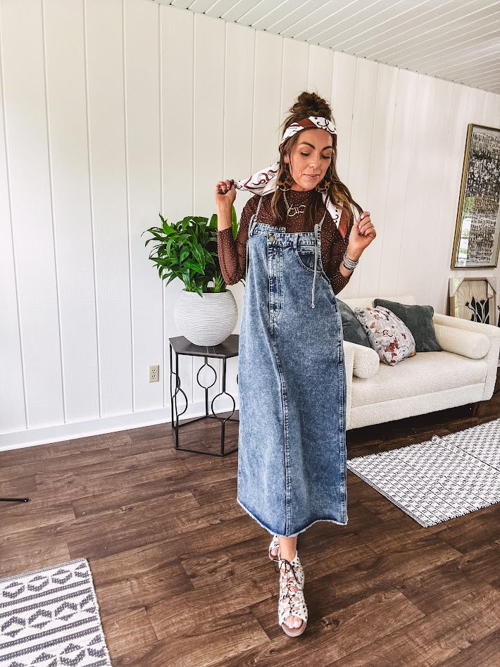Medium wash denim dress with spaghetti straps and silver metal adjusters, a unique button and zipper front resembling the top of a pair of jeans, back pockets, side pockets, a back slit and raw hem.