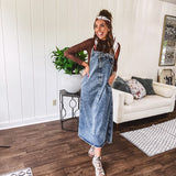 Medium wash denim dress with spaghetti straps and silver metal adjusters, a unique button and zipper front resembling the top of a pair of jeans, back pockets, side pockets, a back slit and raw hem.