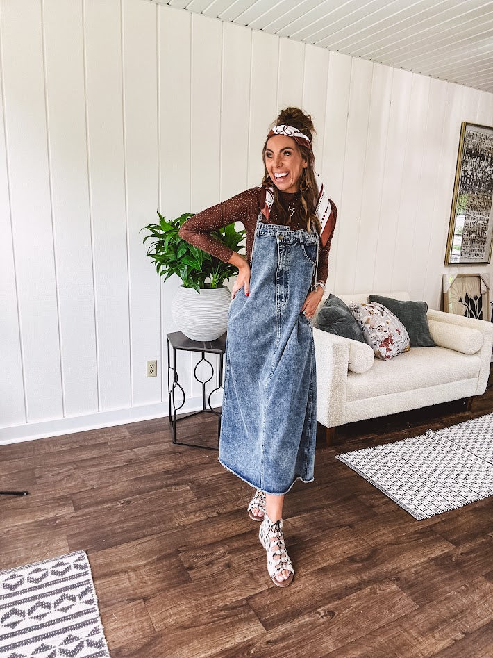Medium wash denim dress with spaghetti straps and silver metal adjusters, a unique button and zipper front resembling the top of a pair of jeans, back pockets, side pockets, a back slit and raw hem.