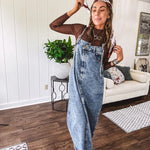 Medium wash denim dress with spaghetti straps and silver metal adjusters, a unique button and zipper front resembling the top of a pair of jeans, back pockets, side pockets, a back slit and raw hem.