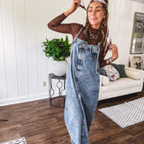 Medium wash denim dress with spaghetti straps and silver metal adjusters, a unique button and zipper front resembling the top of a pair of jeans, back pockets, side pockets, a back slit and raw hem.