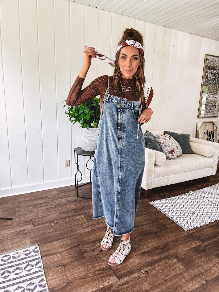 Medium wash denim dress with spaghetti straps and silver metal adjusters, a unique button and zipper front resembling the top of a pair of jeans, back pockets, side pockets, a back slit and raw hem.