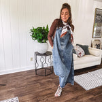 Medium wash denim dress with spaghetti straps and silver metal adjusters, a unique button and zipper front resembling the top of a pair of jeans, back pockets, side pockets, a back slit and raw hem.