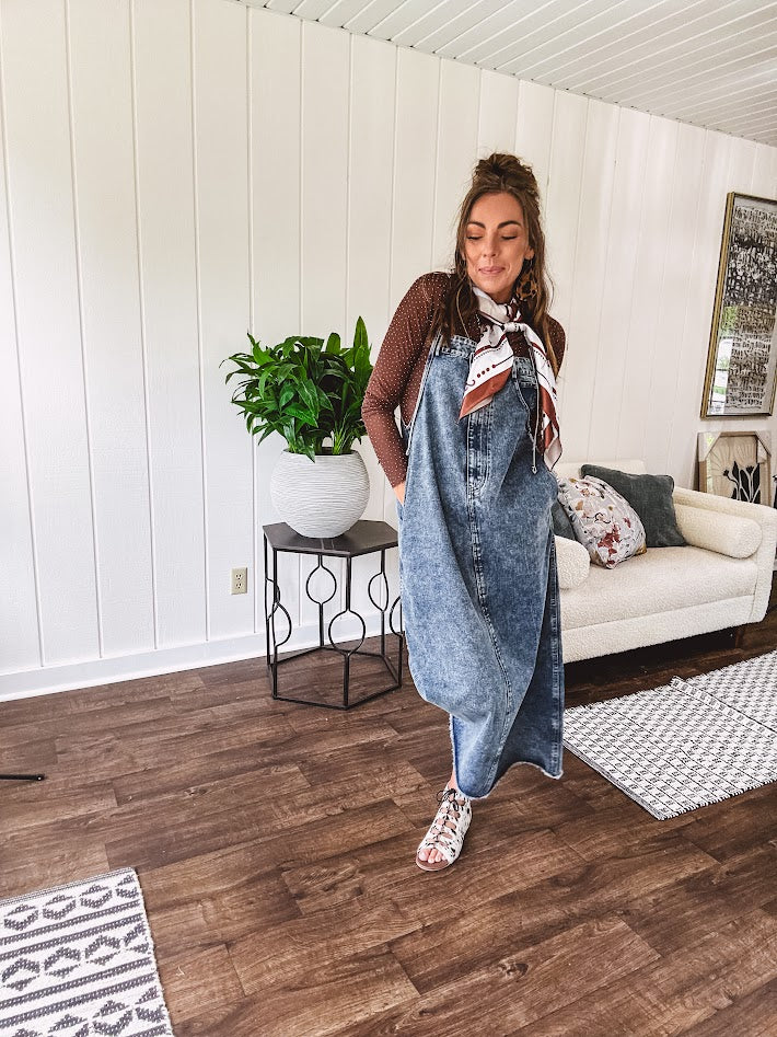 Medium wash denim dress with spaghetti straps and silver metal adjusters, a unique button and zipper front resembling the top of a pair of jeans, back pockets, side pockets, a back slit and raw hem.