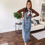 Medium wash denim dress with spaghetti straps and silver metal adjusters, a unique button and zipper front resembling the top of a pair of jeans, back pockets, side pockets, a back slit and raw hem.