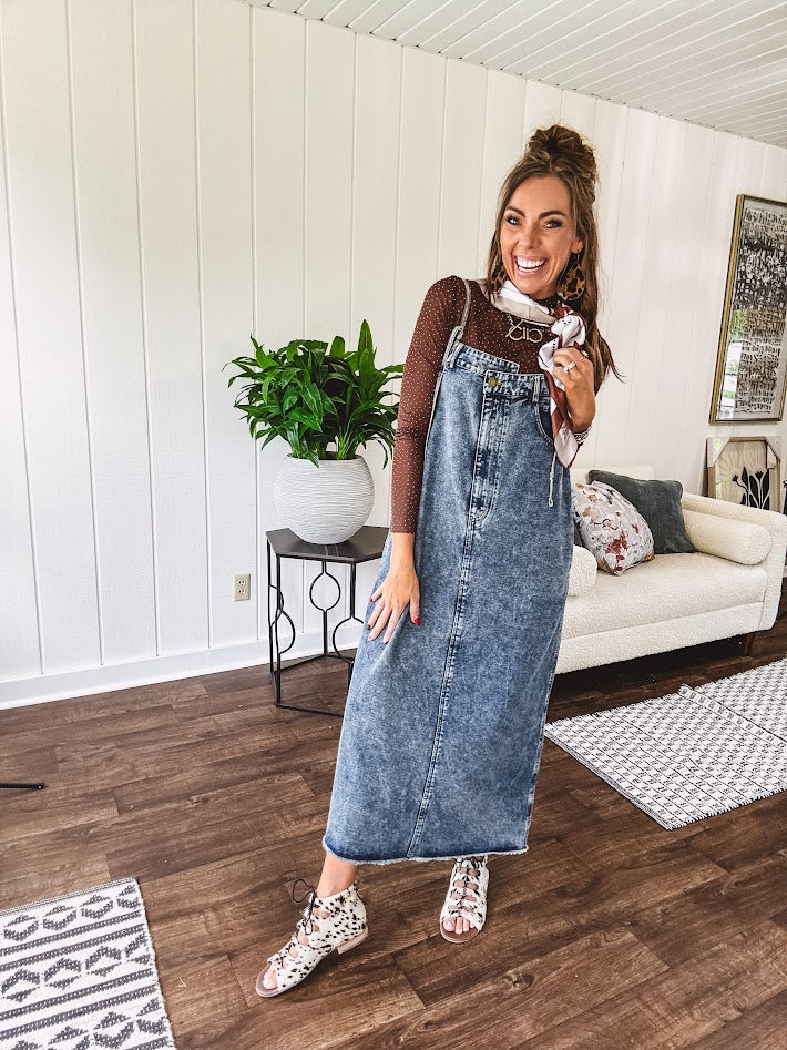Medium wash denim dress with spaghetti straps and silver metal adjusters, a unique button and zipper front resembling the top of a pair of jeans, back pockets, side pockets, a back slit and raw hem.