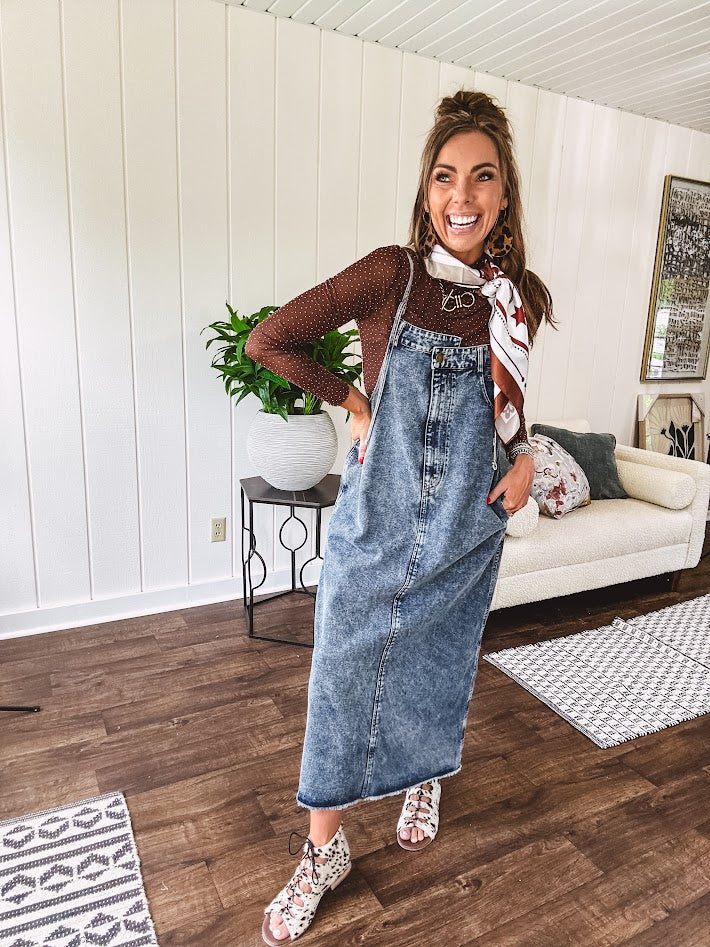 Medium wash denim dress with spaghetti straps and silver metal adjusters, a unique button and zipper front resembling the top of a pair of jeans, back pockets, side pockets, a back slit and raw hem.
