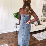 Medium wash denim dress with spaghetti straps and silver metal adjusters, a unique button and zipper front resembling the top of a pair of jeans, back pockets, side pockets, a back slit and raw hem.