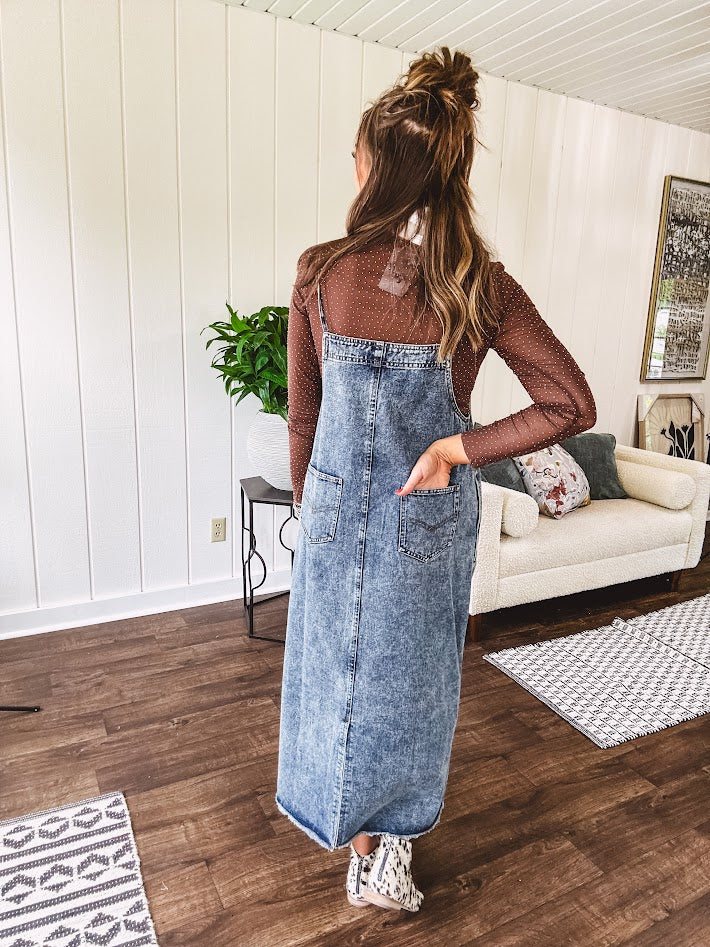Medium wash denim dress with spaghetti straps and silver metal adjusters, a unique button and zipper front resembling the top of a pair of jeans, back pockets, side pockets, a back slit and raw hem.