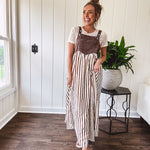 Brown washed denim overall top with silver metal hardware and adjustable straps and a cream and brown striped, lightweight skirt with pockets.