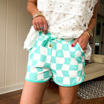 Silky athleisure style shorts with elastic drawstring waist with gold accents, pockets in a teal and white checkered pattern with teal trim.