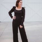 Black silky jumpsuit with puffed long sleeves, square neckline, side cutouts, elastic at the back waist and a tie at the back neckline.