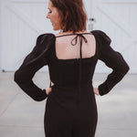 Black silky jumpsuit with puffed long sleeves, square neckline, side cutouts, elastic at the back waist and a tie at the back neckline.