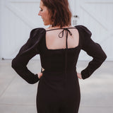 Black silky jumpsuit with puffed long sleeves, square neckline, side cutouts, elastic at the back waist and a tie at the back neckline.